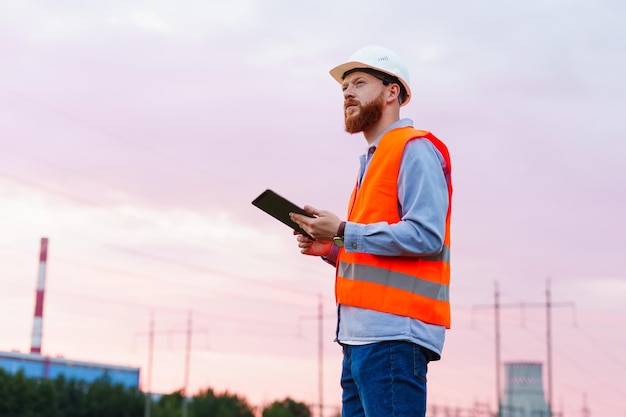 An engineer with a tablet on the background of a power plant Inspection of engineering systems A man in a helmet and an orange vest at sunset