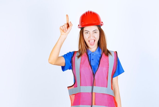 Foto donna ingegnere in uniforme e casco rosso che mostra qualcosa.