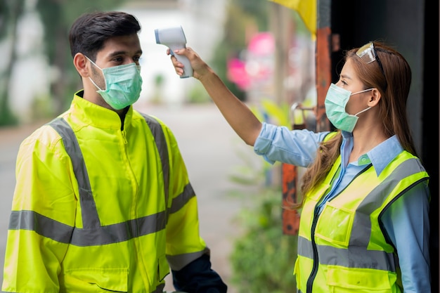 Engineer worker waring surgical mask checking body temperature using infrared digital thermometer check temperature before into work place, workers with protective mask