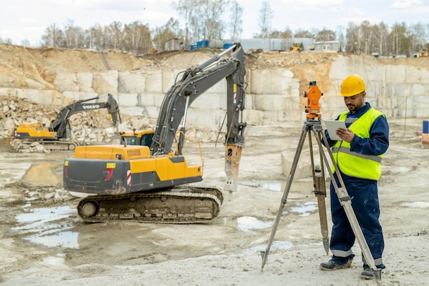 Engineer in workwear standing by geodetic station against bulldozer