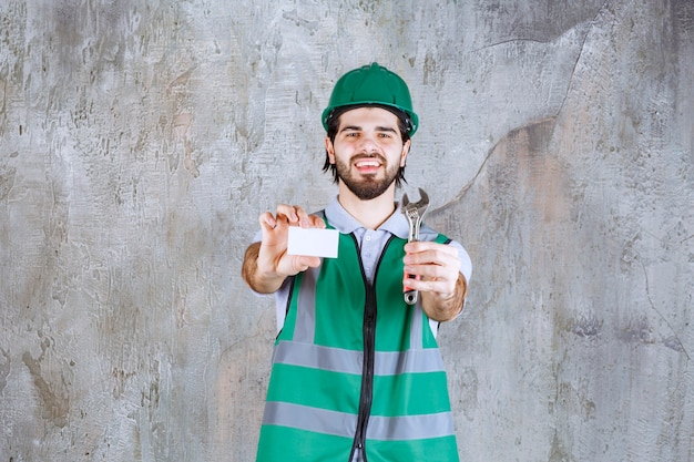 Engineer in yellow gear and helmet holding a metallic wrench and presenting his business card. 