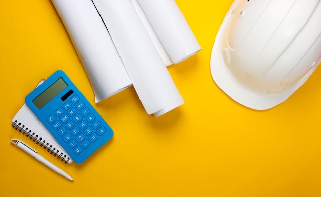 Engineering construction safety helmet, blueprint rolls and calculator, notebook on yellow. Calculation of the cost building a house. Flat lay