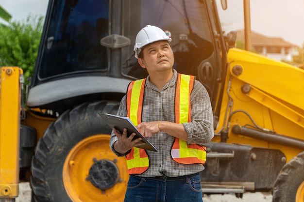 Photo engineering wearing a white safety helmet standing in front of the backhoe looking at home construction work