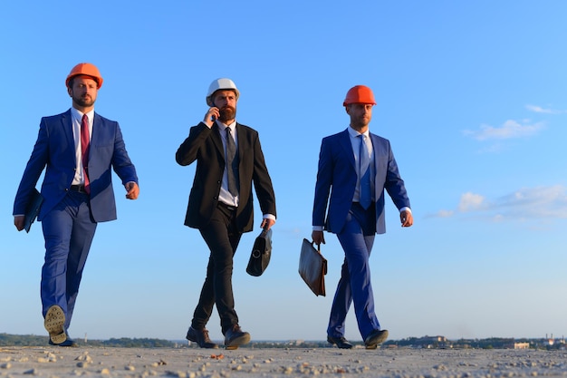 Engineers hold black and brown cases. Architects with strict faces in suits, white and orange helmets. Builders walk confidently and talk on phone on blue sky background. Business and building concept