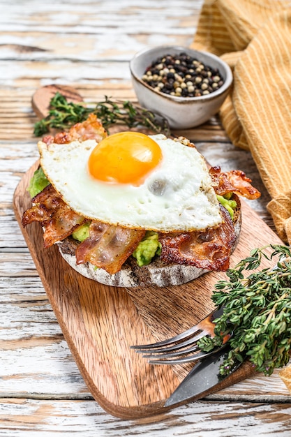 English breakfast, toast with bacon, avocado and egg on a cutting board. Healthy food.  White wooden background. Top view.