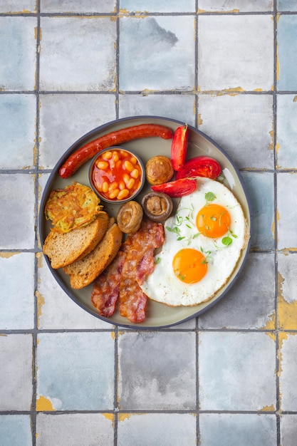 Colazione inglese con uova fritte salsicce pancetta fagioli toast pomodori e funghi sfondo piastrelle...