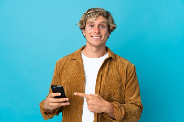 Англичанин над изолированной синей стеной, используя мобильный телефон и указывая на него