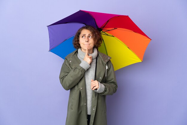 English woman holding an umbrella isolated on purple background having doubts and thinking