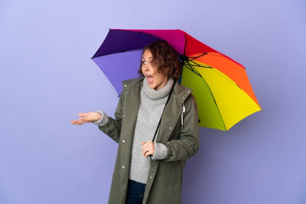 English woman holding an umbrella isolated on purple background with surprise expression while looking side