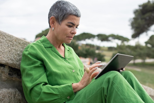 Donna senior assorta che legge il libro elettronico nel parco. modello femminile con capelli grigi corti in abiti luminosi che tengono tablet. tempo libero, concetto di letteratura