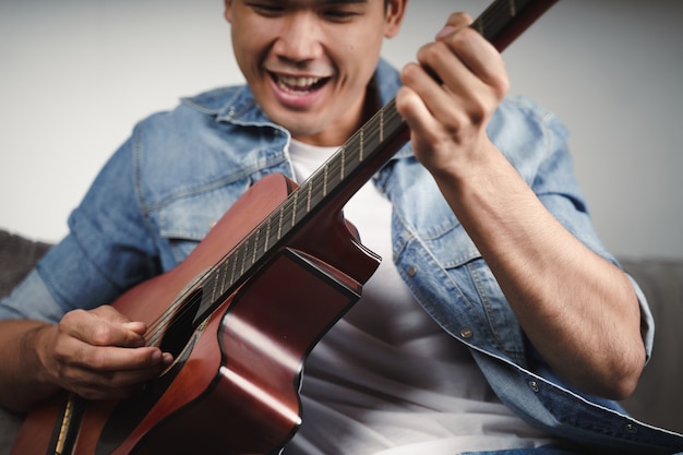Наслаждайтесь красивым азиатским мужчиной, который тренируется или играет на гитаре на диване в гостиной
