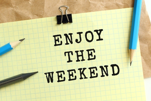 enjoy the weekend lettering on a yellow sheet of paper over crumpled kraft paper. pencils and paper clip.
