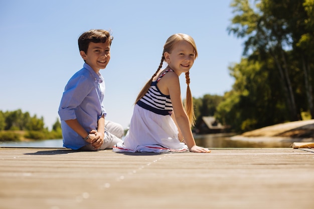 Enjoyable day. Upbeat little siblings posing for the camera and smiling while sitting on a pier and basking in the sunlight