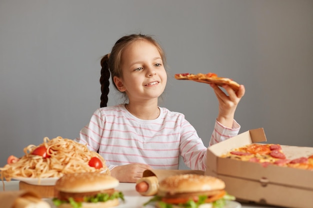 Фото Приятная улыбающаяся красивая маленькая девочка с косами сидит за столом с нездоровой пищей, изолированной на сером фоне, держа большой кусок пиццы и читая, чтобы съесть нездоровое блюдо