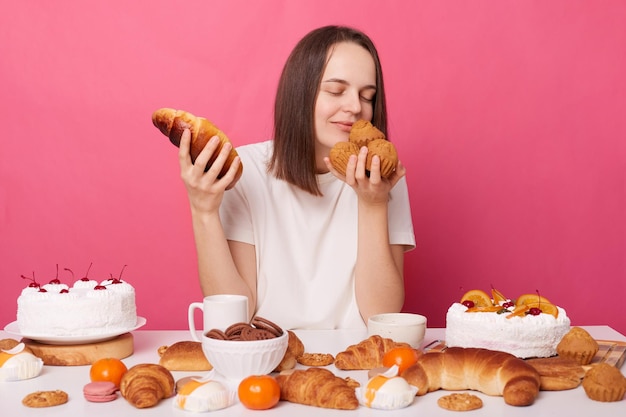 흰색 티셔츠를 입고 축제 테이블에 앉아 분홍색 배경에서 격리된 맛있는 페이스트리 냄새가 나는 홈메이드 과자를 들고 있는 즐거운 여성