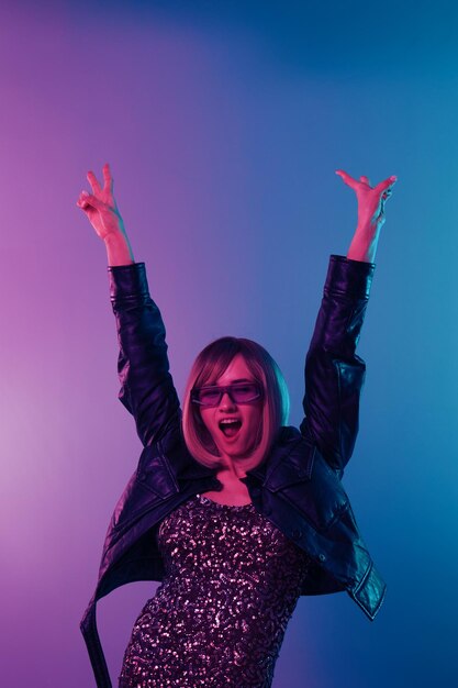 Photo enjoyed sexy beautiful blonde woman in leather jacket sparkly dress trendy sunglasses dance hold hands up show vsign posing isolated in blue pink color light studio background neon party concept