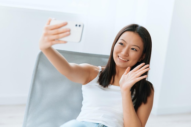 Enjoyed smiling tanned lovely young asian woman waving hand\
greeting friend on live doing selfie video call at home distance\
communication social media concept cool offer banner wide\
angle