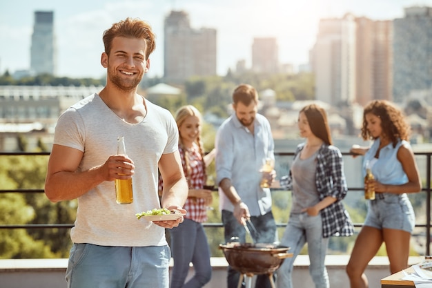 Godersi il barbecue con gli amici, un uomo giovane e allegro tiene in mano una bottiglia di birra e un piatto