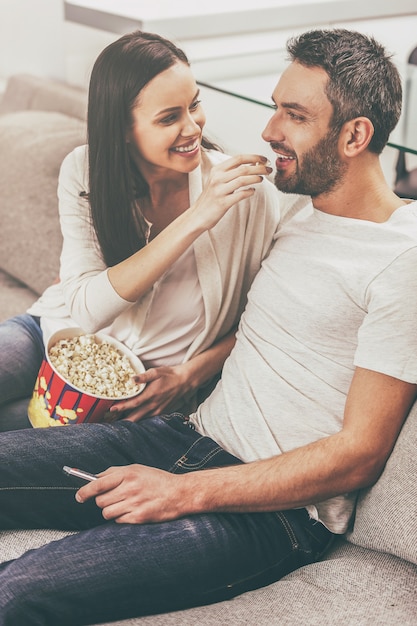 Наслаждаемся беззаботным временем вместе. Красивая молодая влюбленная пара привязывается друг к другу и ест попкорн, сидя на диване и смотря телевизор