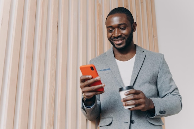 Photo enjoying coffee break at work young positive african man office worker in suit using mobile phone