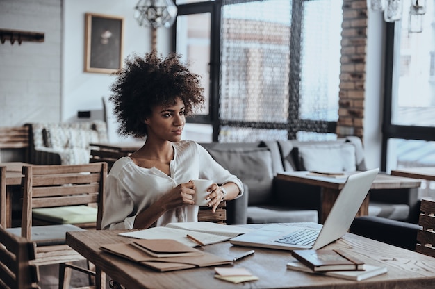 Enjoying coffee at her working place. Beautiful young African woman holding a cup and looking away 