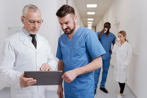 Godersi la differenza tra le generazioni. pediatri gioiosi intelligenti alla mano che godono del lavoro in ospedale e lavorano con il tablet mentre altri colleghi hanno una pausa caffè in background