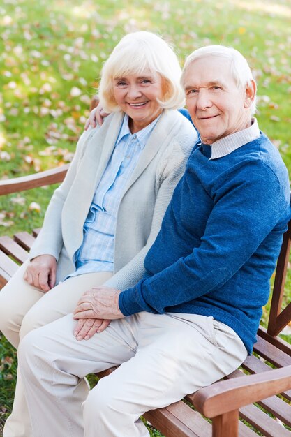 Enjoying each other. Top view of happy senior couple holding hands and looking at camera with smile while sitting on the park bench together