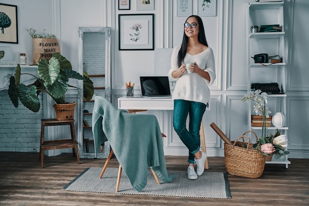 Enjoying fresh coffee. Full length of beautiful young woman looking away and smiling while standing in home office