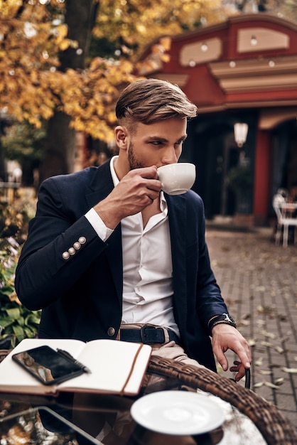 Enjoying fresh coffee. Handsome young man in smart casual wear drinking coffee while sitting in restaurant outdoors