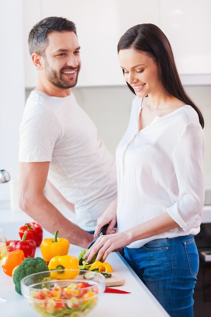 Enjoying healthy lifestyle. Beautiful young and cheerful woman cutting vegetable in the kitchen while her husband standing close to her and smiling