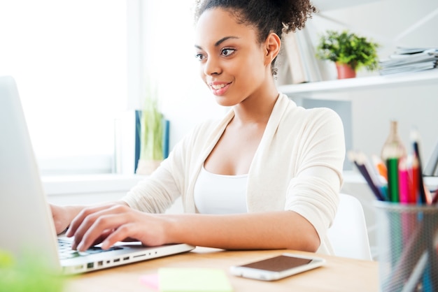 Enjoying her work in office. Beautiful young African woman working at the laptop and smiling while sitting at her working place in office