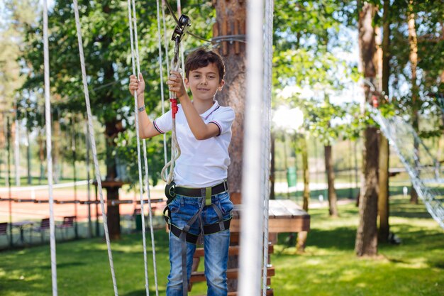 Наслаждаясь собой. Радостный малолетний мальчик веселится в веревочном парке и приятно улыбается, перемещаясь на качелях