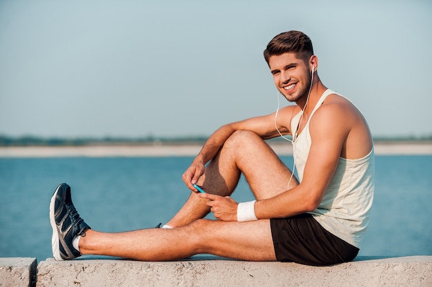 Enjoying his favorite music. Side view of cheerful young muscular man in headphones holding MP3 player and smiling while sitting on parapet