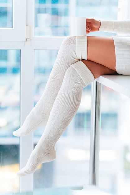 Photo enjoying hot drink at home. close-up of woman in white warm socks holding coffee cup while sitting in front of the window at home