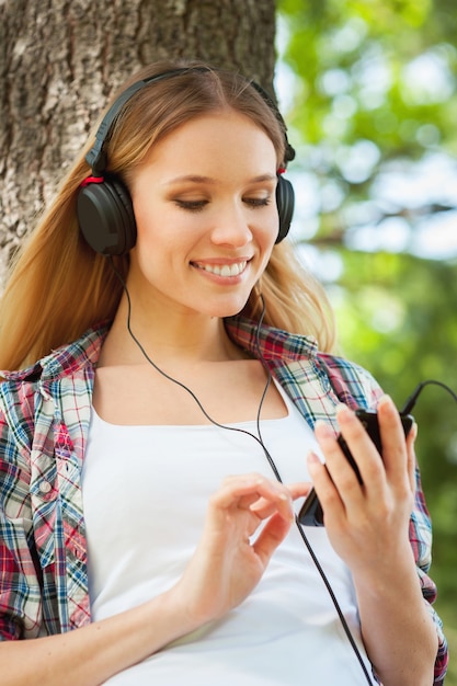 Enjoying music and fresh air. Beautiful young woman in headphones listening to the music and smiling while leaning at the tree in a park