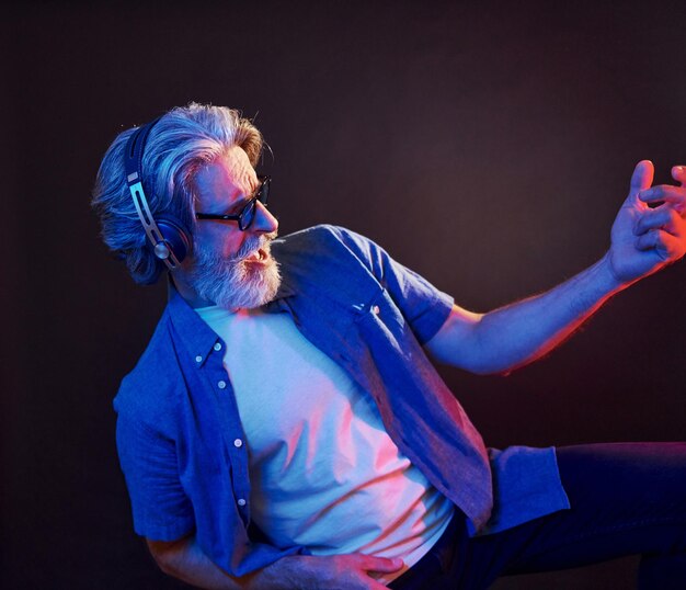 Enjoying music Neon lighting Stylish modern senior man with gray hair and beard is indoors