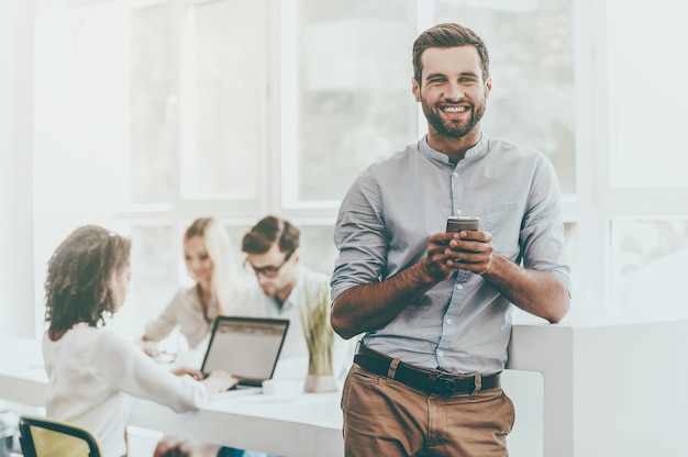 Enjoying office life. Joyful young man holding mobile phone and looking at camera while his colleagues working in the background