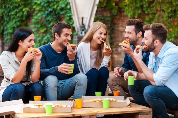 Enjoying pizza together. Group of happy young people eating pizza while sitting outdoors