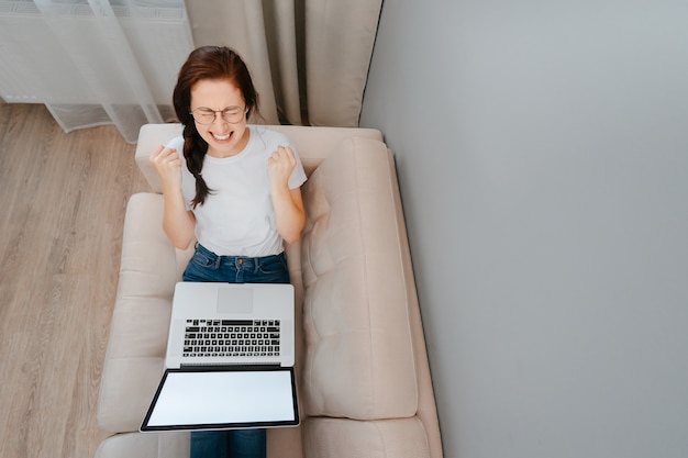 Enjoying the rest at home young smiling woman is working on a laptop on a large comfortable sofa and...