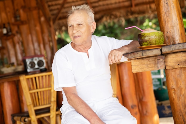 Enjoying the retirement. Happy senior man is drinking a coconut water in the beach bar.