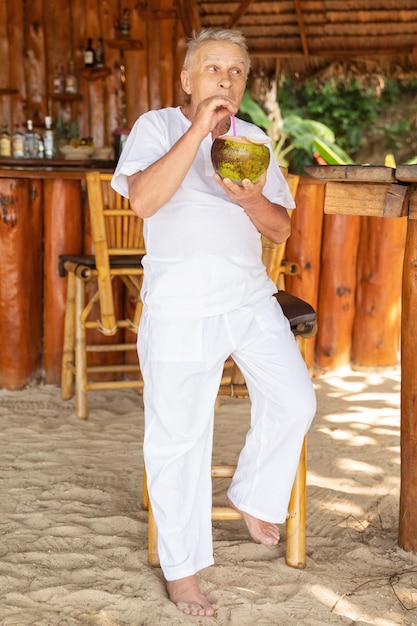 Enjoying the retirement. Happy senior man is drinking a coconut water in the beach bar.