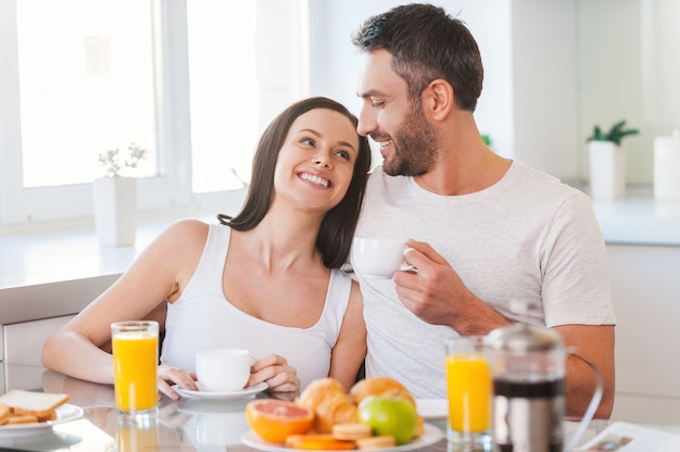 Наслаждаемся воскресным утром вместе. Красивая молодая пара обнимаются и улыбаются, сидя на кухне вместе и завтракая