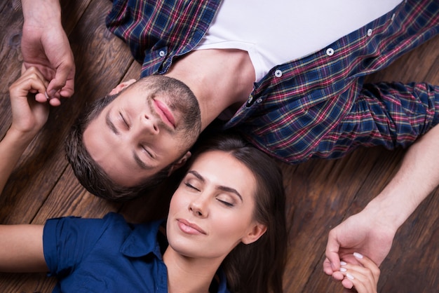 Enjoying their closeness. Top view of beautiful young loving couple holding hands and keeping eyes closed while lying together on the hardwood floor