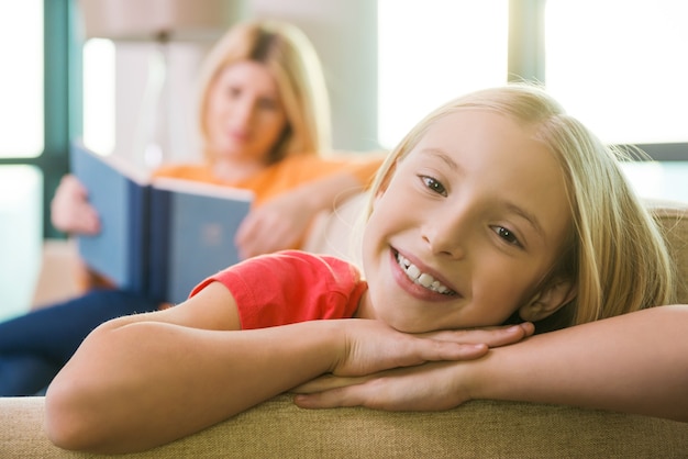 Enjoying time near mother. happy little girl sitting on the couch and smiling while her mother reading book in the background