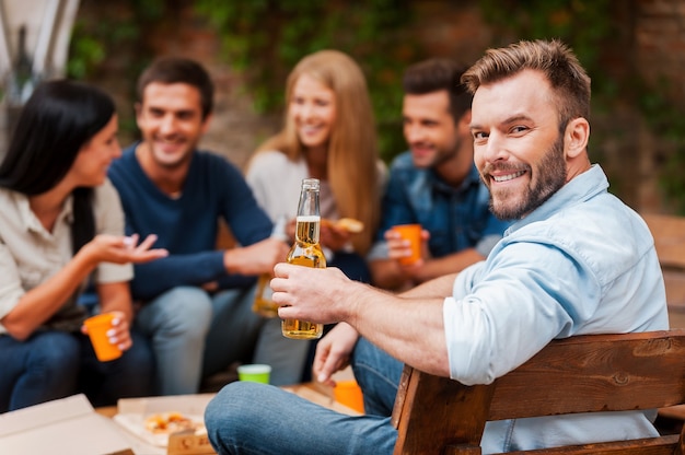 Enjoying time with friends. Happy young man holding bottle with beer and looking at camera while his friends talking to each other in the background