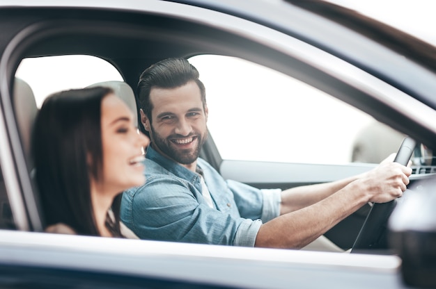 Enjoying the trip.  Young couple sitting in the car and smiling while handsome man holding hands on the steering wheel