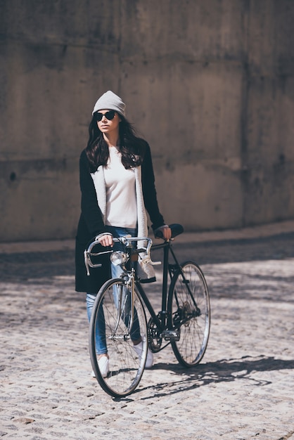 Наслаждаемся городским образом жизни. Полная длина красивой молодой женщины в солнцезащитных очках катит велосипед и смотрит в сторону во время прогулки на свежем воздухе