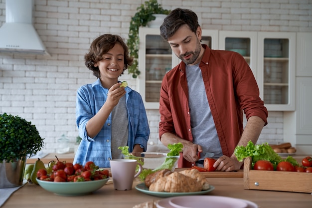 Godendo il fine settimana a casa giovane papà e figlio felice che tagliano verdure fresche preparando un'insalata