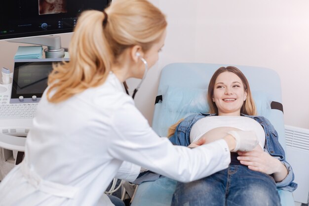 Enjoying working with future mothers. Involved qualified mature obstetrician working in the clinic while checking pregnant belly and using stethoscope for detecting heat beating