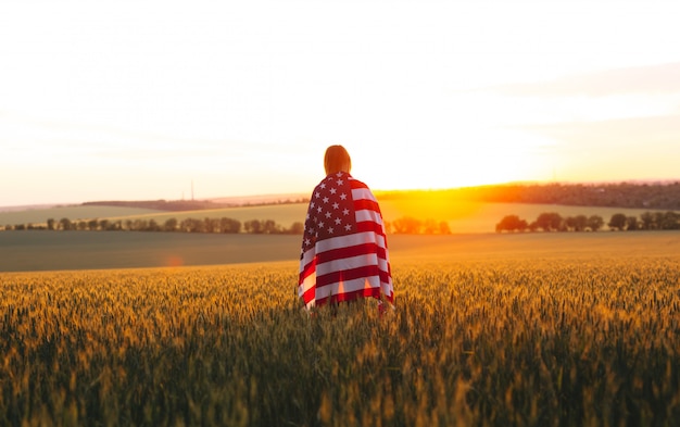 夕暮れ時の麦畑でアメリカの国旗を持つenjoyng女性。 7月4日。独立記念日、愛国的な休日。